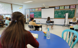 A teacher sitting at a desk behind students at their desks