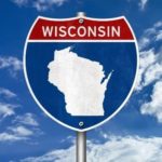 Text contains Wisconsin. Image contains shape of Wisconsin on a road sign with sky and clouds behind.
