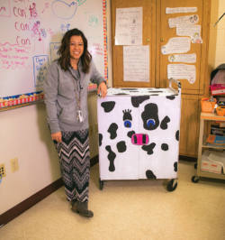 Images shows a teacher next to a computer charging cart on wheels painted like a cow.